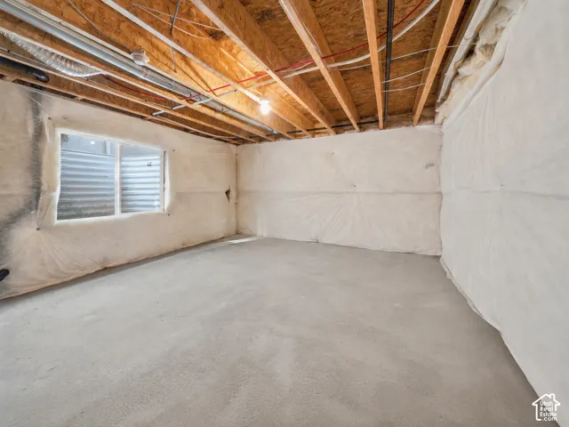 Unfinished lower level featuring exposed wood-joist ceiling, concrete flooring, foundation walls covered in protective vapor barrier, and an egress window with a corrugated steel window well