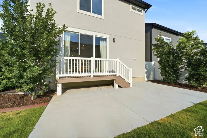 Modern two-story residence featuring a spacious concrete patio, white composite deck railing with glass panels, mature landscaping with established trees and shrubs, and contemporary architectural styling with mixed exterior finishes including taupe sidin