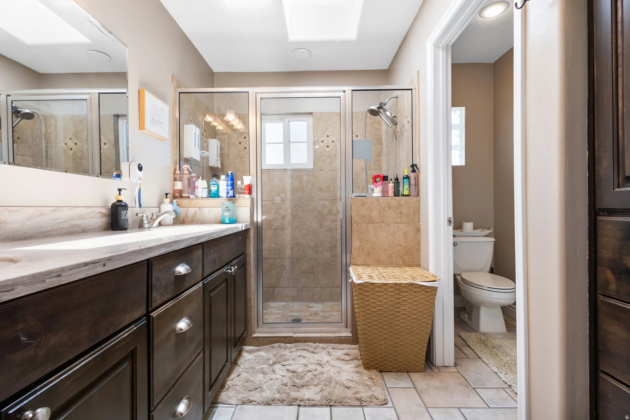 Dark wood-finish vanity with a light-toned countertop, glass-enclosed shower with tiled surround, recessed lighting, and tile flooring