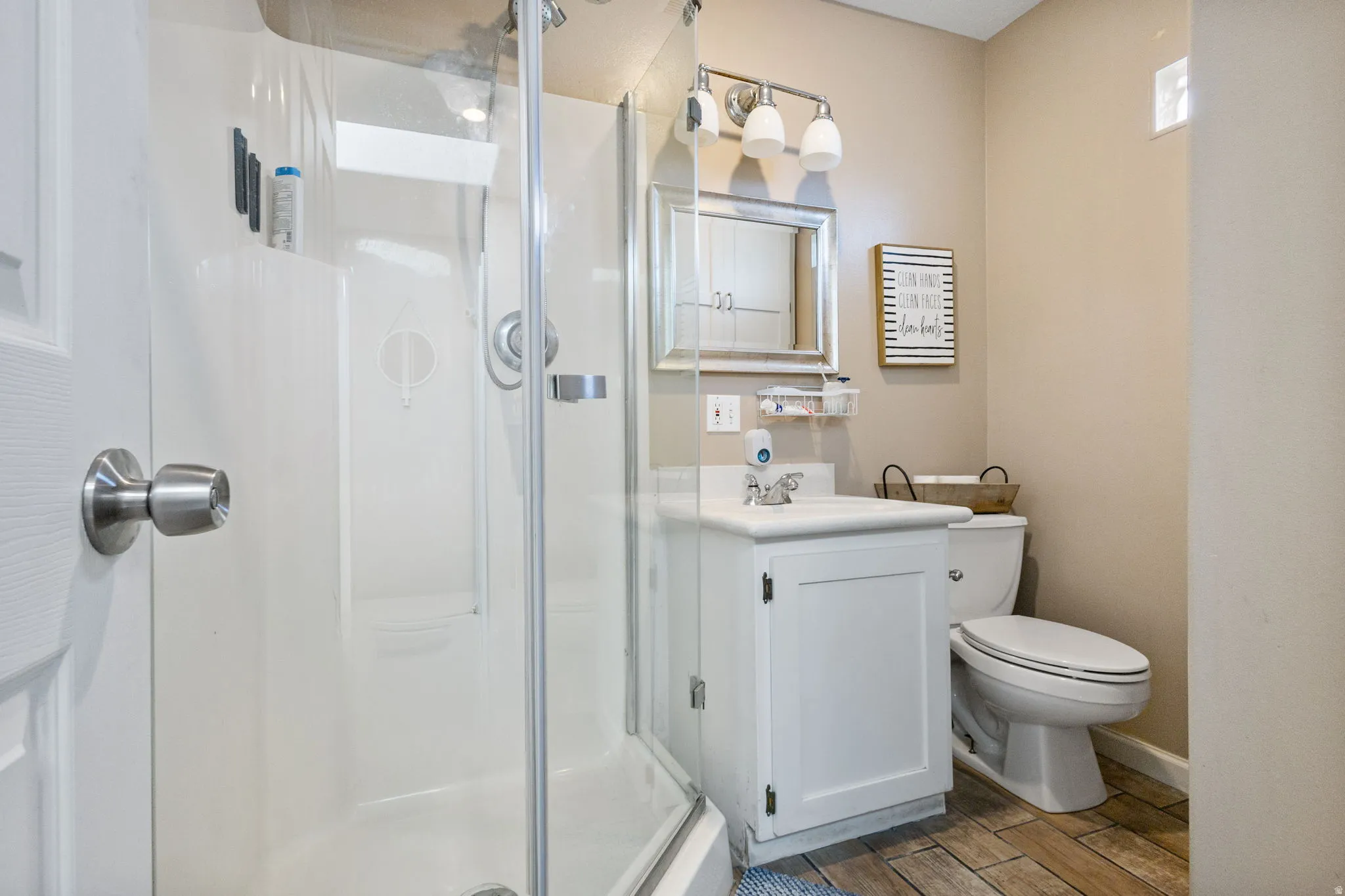 Glass-enclosed shower with chrome fixtures, single vanity with white cabinetry, wall-mounted mirror, and wood-finish flooring