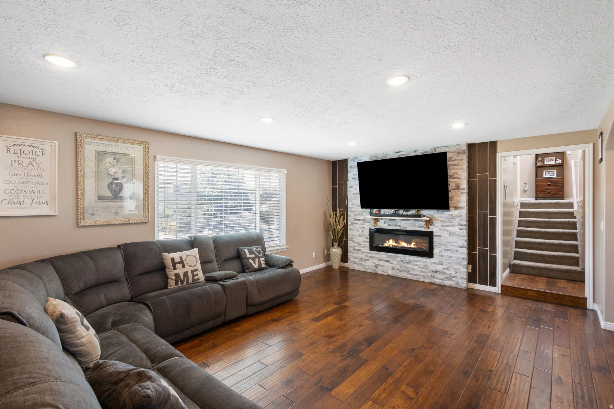 Hardwood-finish flooring, recessed lighting, white window blinds, stone-clad fireplace with mantel, and carpeted stairs