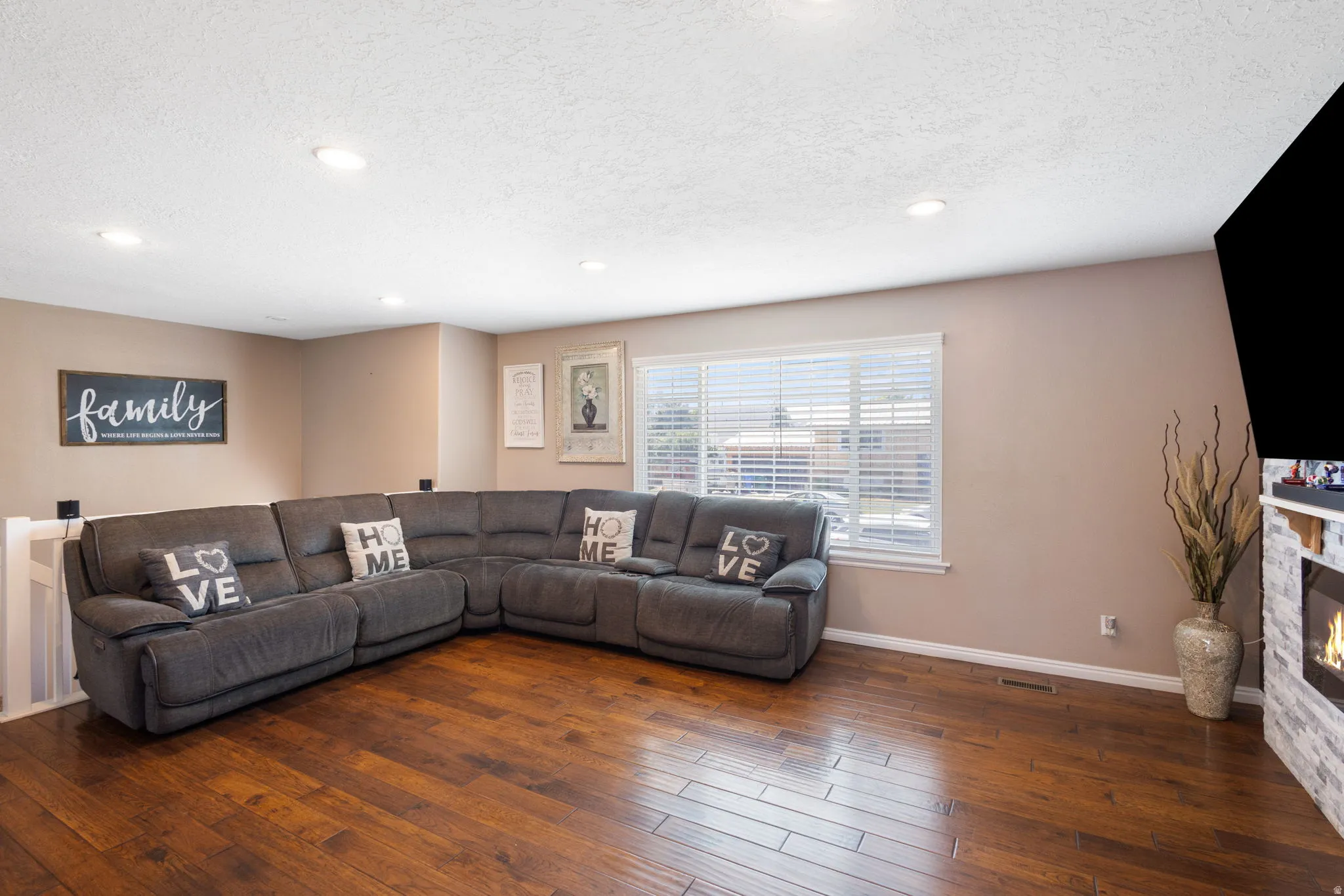 Hardwood flooring, recessed lighting, white window blinds, light neutral wall paint, and a built-in stone-clad fireplace