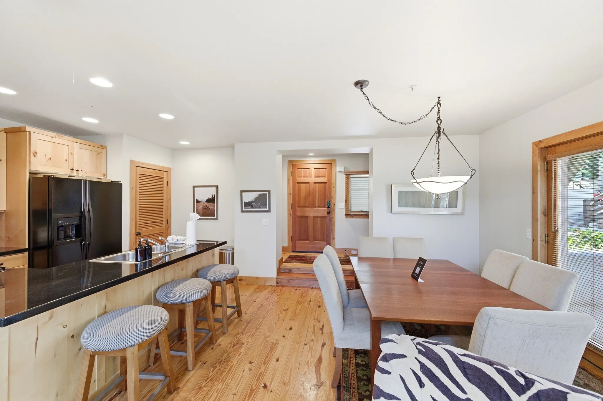 Open-concept kitchen and dining area featuring wood-finish flooring throughout, custom wood cabinetry with black stainless steel appliances, a multi-seat island with wood-frame seating, and a spacious dining table anchored by a pendant light fixture