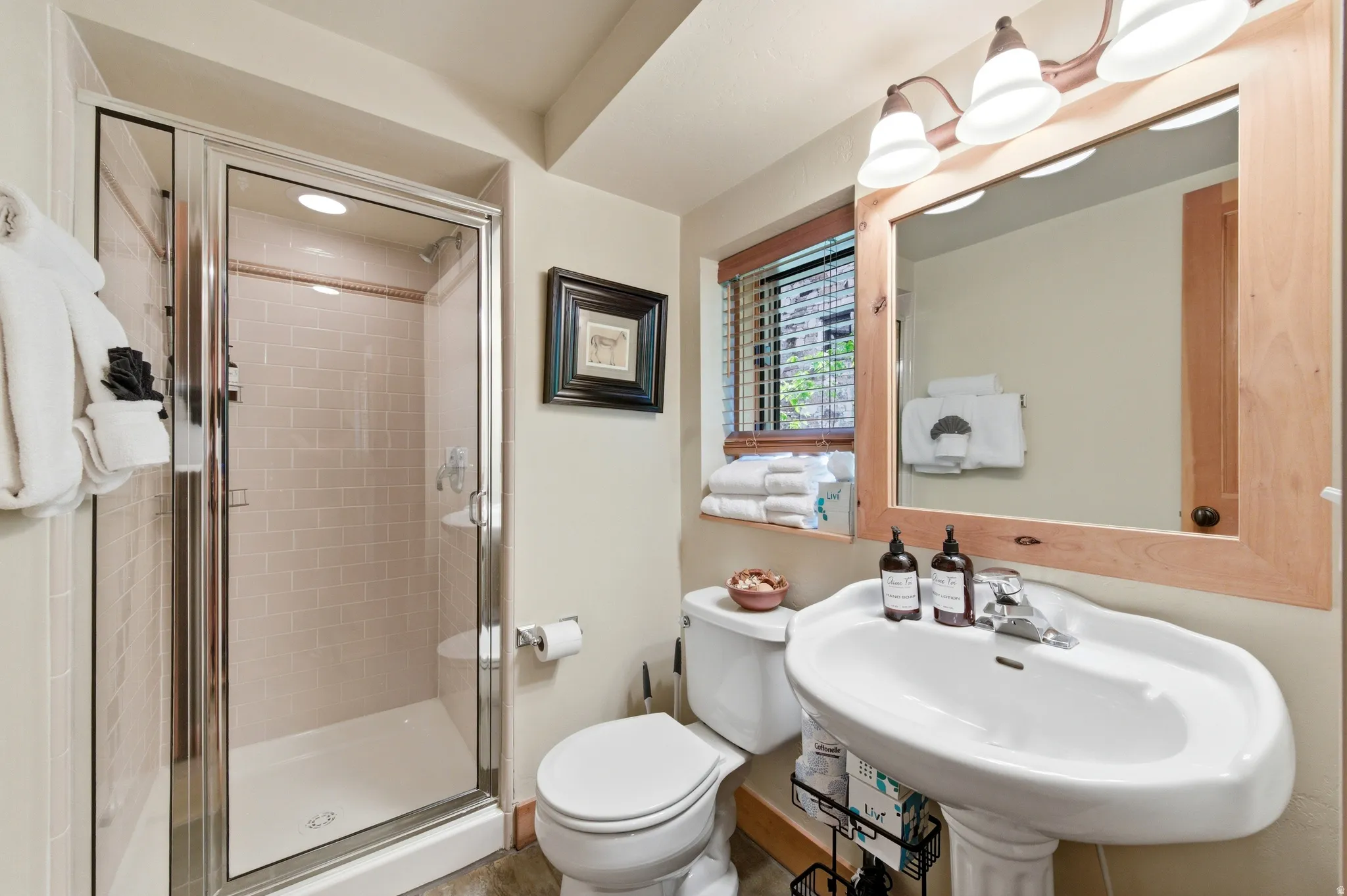Pedestal sink, wood-finish mirror frame, walk-in shower with tile surround, window with blinds