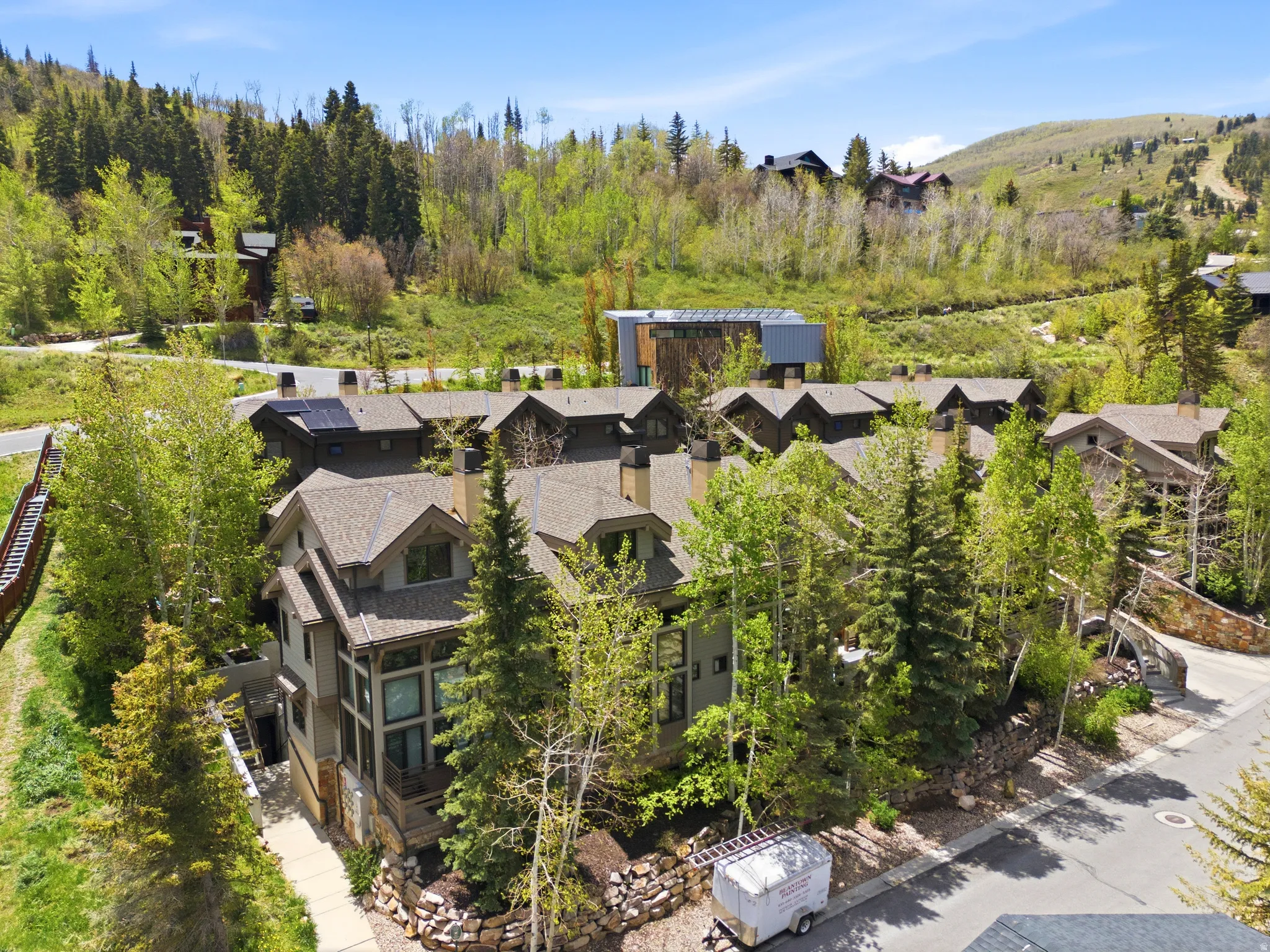 Luxury mountain estate nestled among mature evergreens and aspens, featuring multiple stone and timber-clad residences with peaked rooflines, expansive grounds with natural landscaping, and panoramic views of forested hillsides and rolling terrain