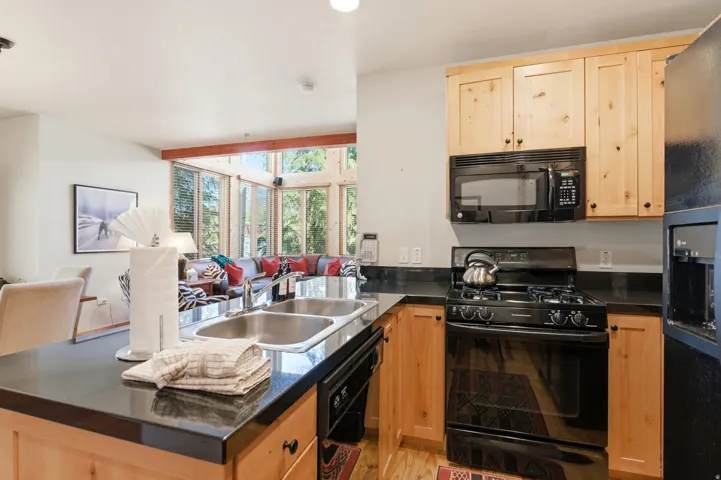 Kitchen featuring light wood cabinetry, dark countertops, a double basin stainless steel sink, and a black appliance suite