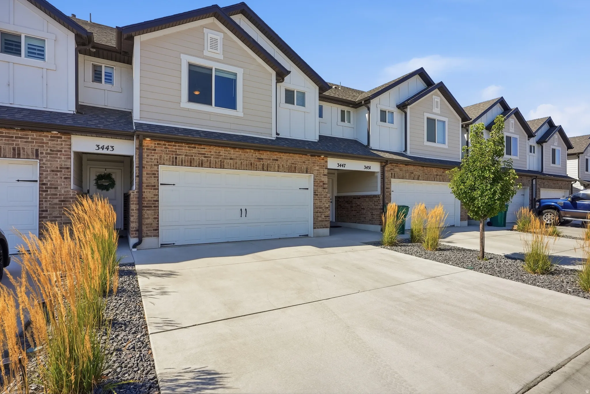 Modern townhome featuring a spacious concrete driveway with integrated garage parking, crisp white and brick exterior with peaked rooflines, decorative landscape edging with ornamental grasses, and a two-car garage with white paneled doors