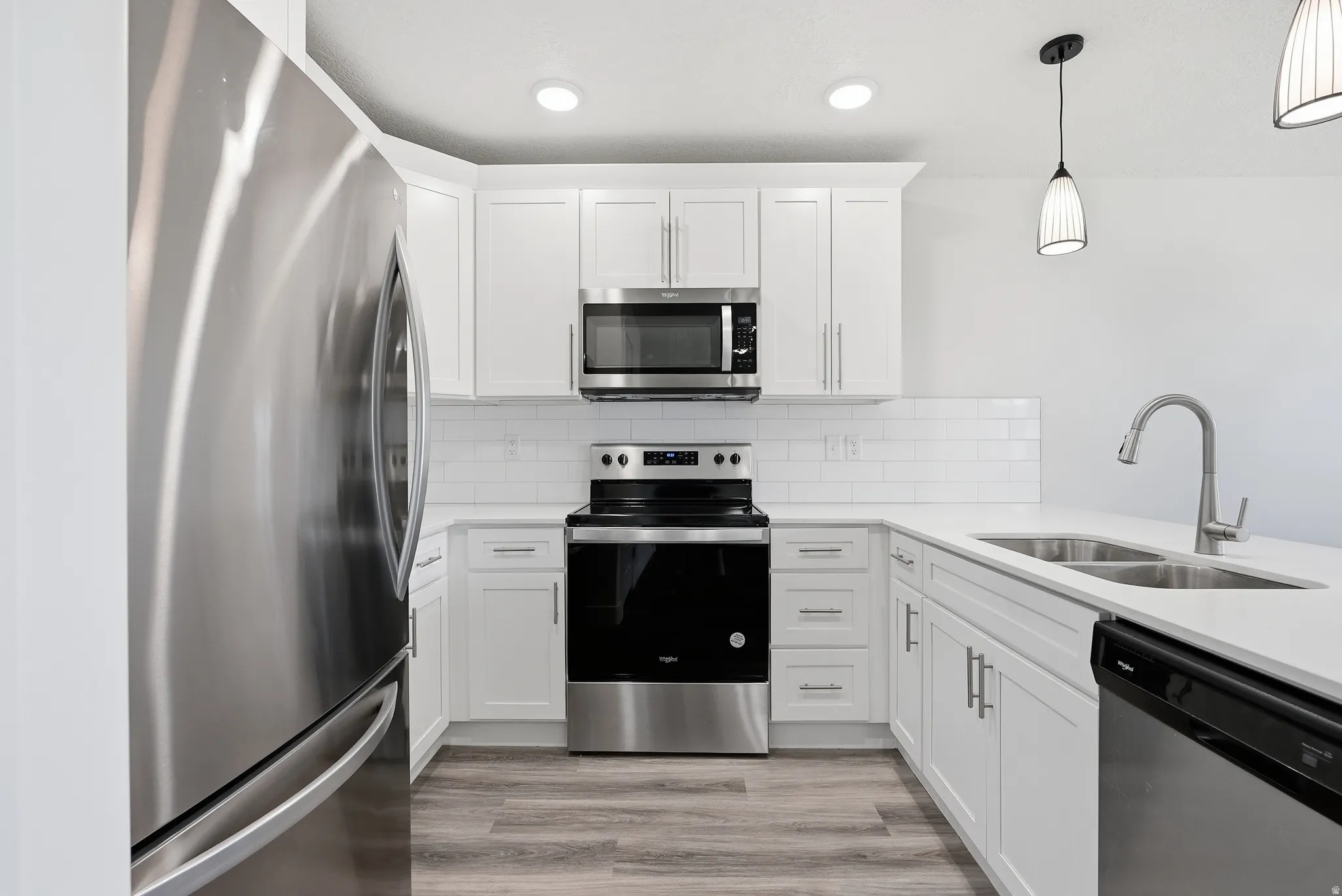 White cabinetry with brushed nickel hardware, stainless steel appliances, white subway tile backsplash, light-toned countertops, and wood-finish flooring