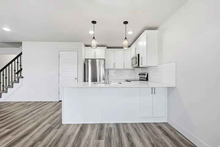 Kitchen island with integrated sink, stainless steel appliances, white subway tile backsplash, white shaker cabinetry, and wood-finish flooring