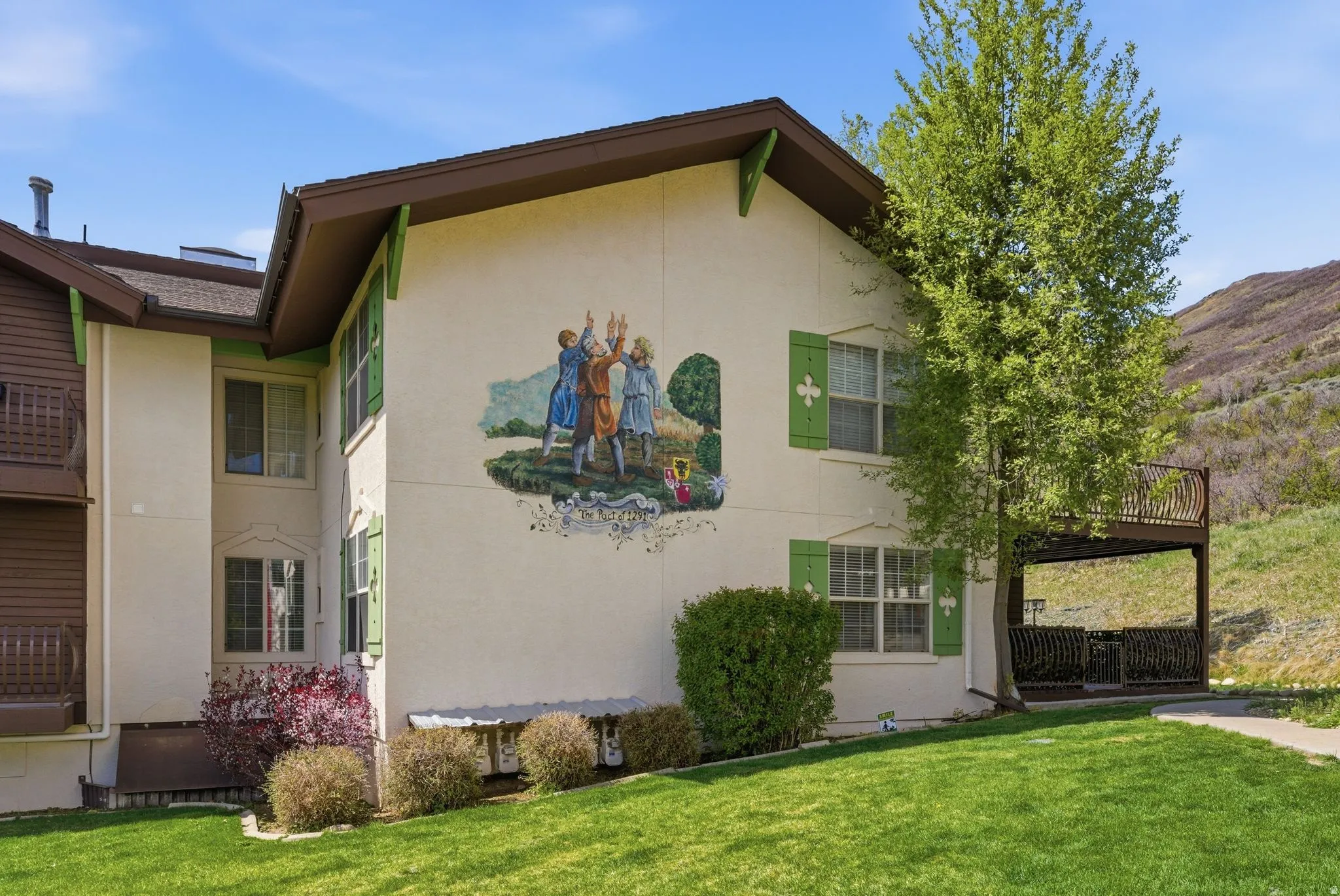 Exterior building featuring a decorative wall mural, multiple windows with green shutters, a brown overhanging roof, and a private upper-level balcony