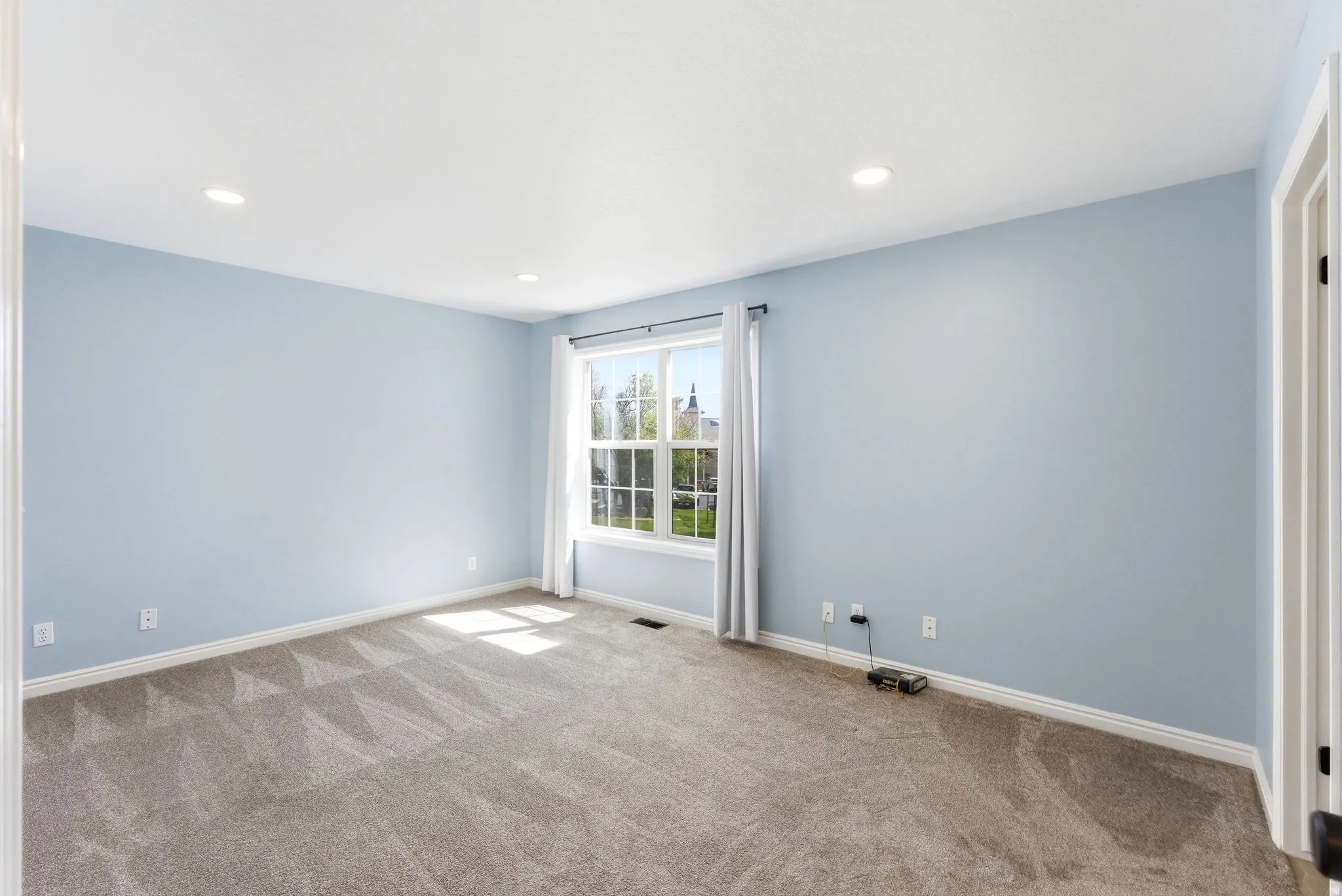Spacious room featuring light blue walls, gray patterned carpeting, recessed lighting, and a white-trimmed window
