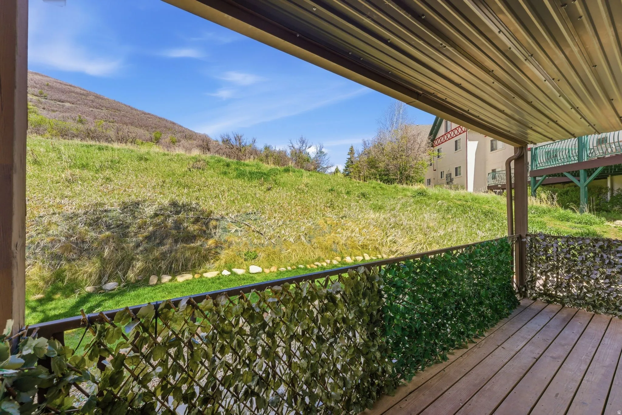 Covered outdoor deck with wood-finish flooring, featuring a decorative privacy screen, metal railing, and an expansive grassy hillside backdrop