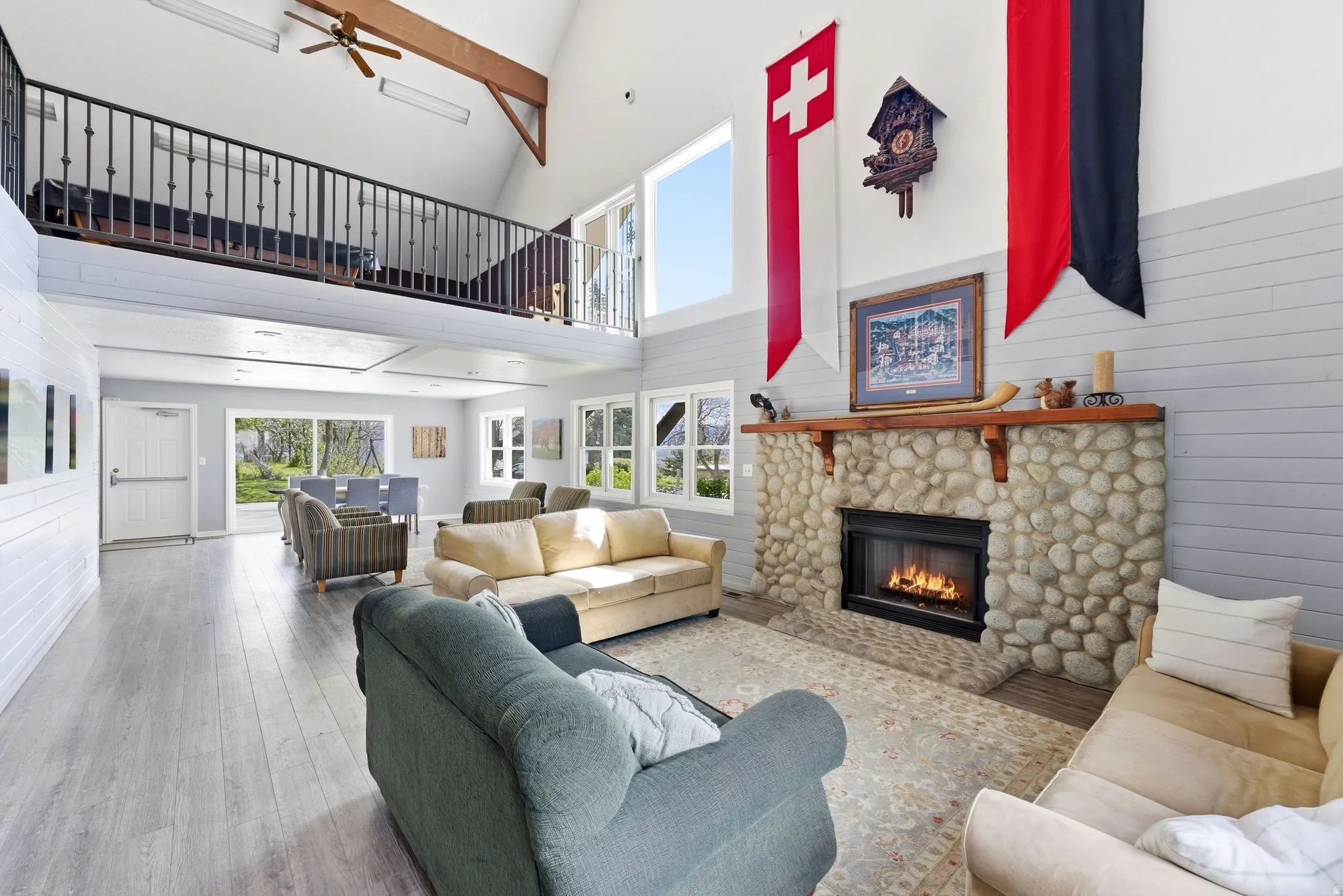 Spacious living area featuring a prominent stone fireplace with a wood mantle, vaulted ceilings with exposed beams, and a second-story balcony with metal railings