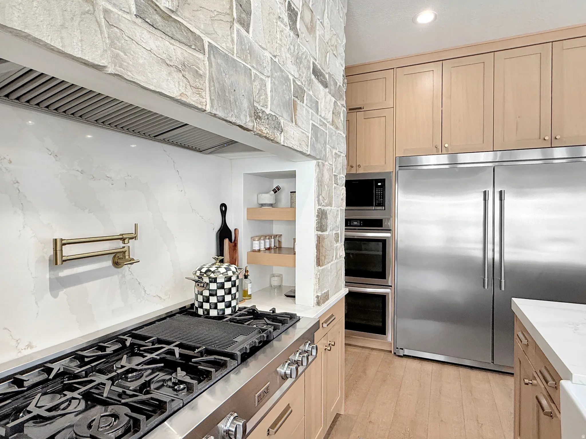 Stone accent wall, stainless steel range, wall-mounted pot filler, light wood-finish cabinetry, built-in stainless steel appliances