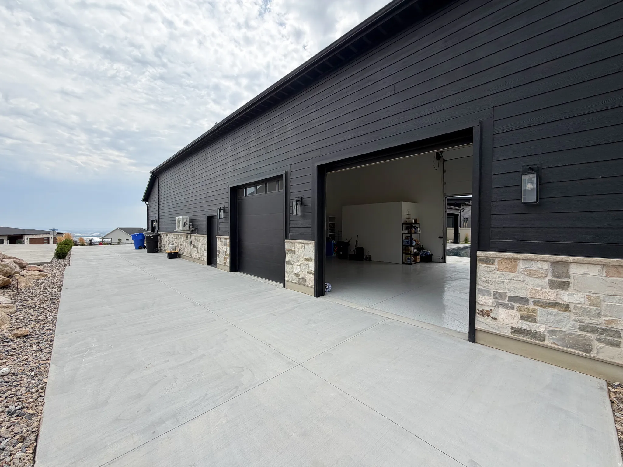 Exterior featuring black horizontal siding, stone veneer accents, two garage doors, and concrete paving