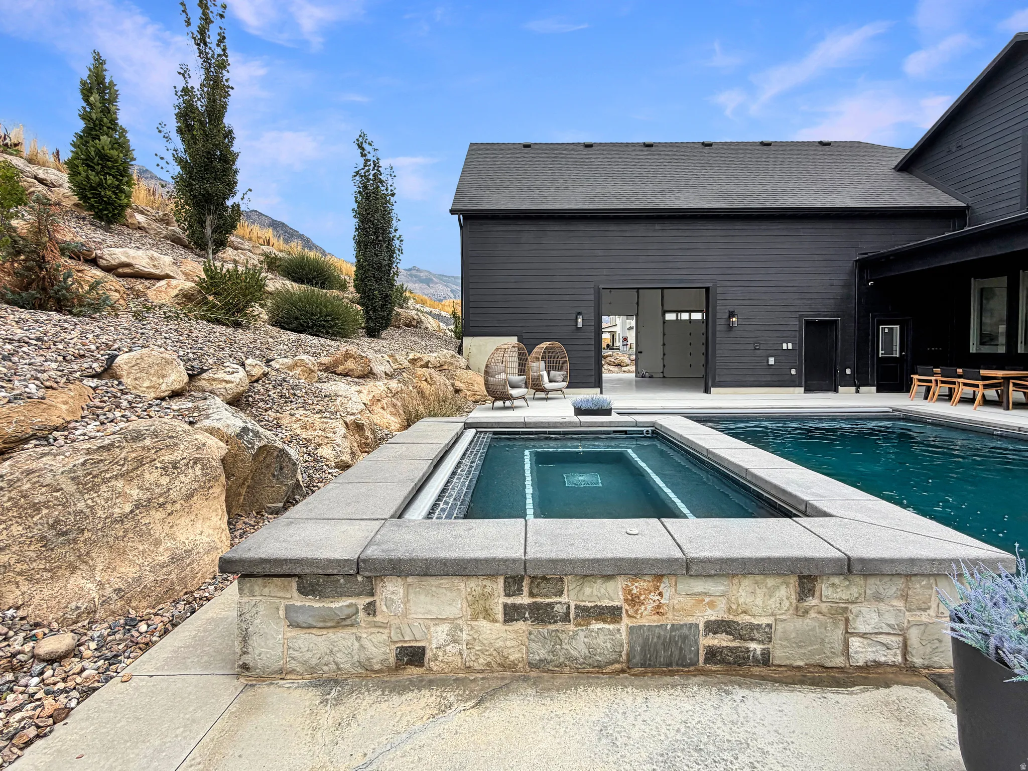 In-ground pool with an integrated spa, stone-clad spa surround, expansive concrete patio, and a dark-toned exterior