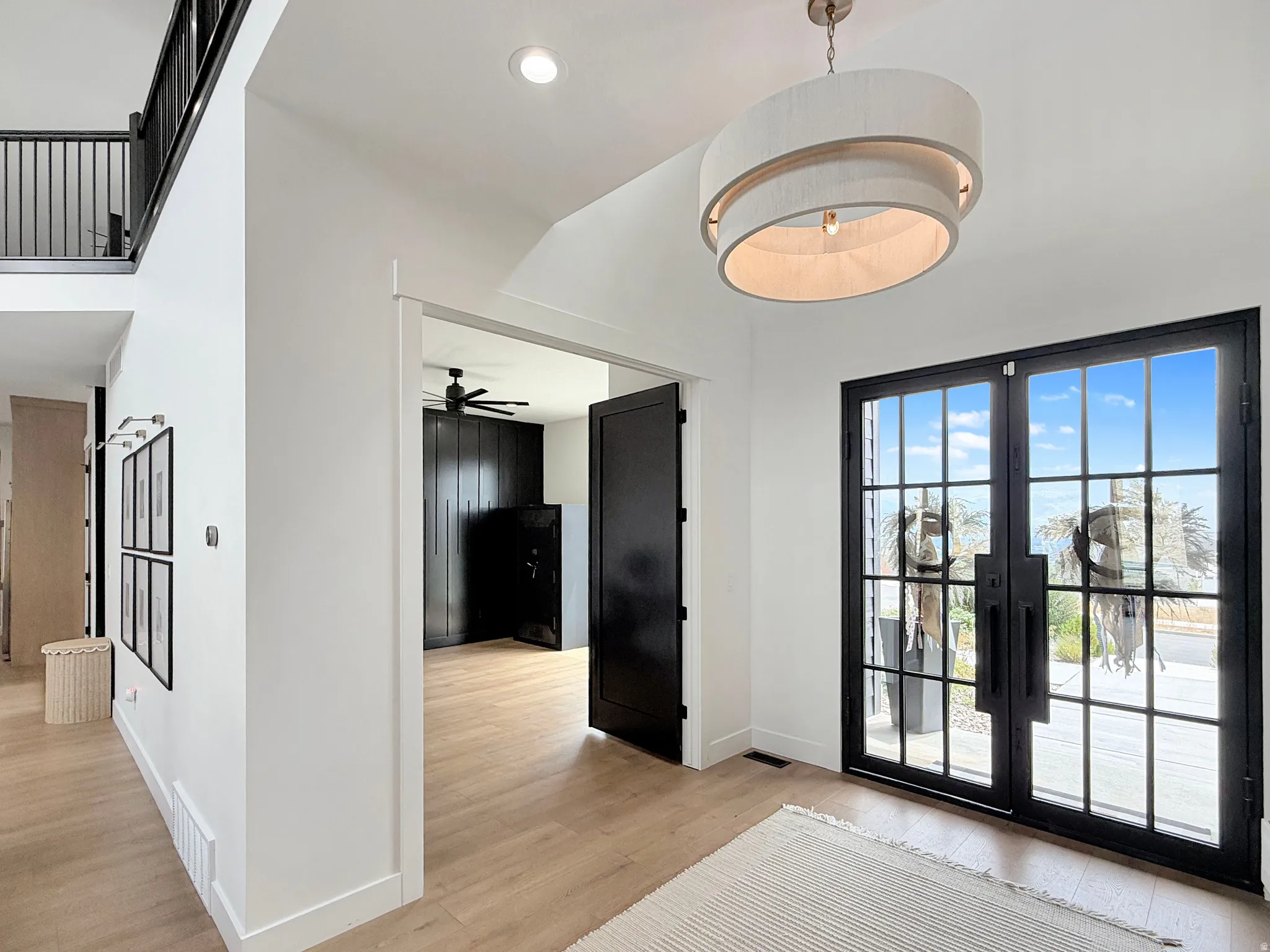 Double entry doors with black frames and glass panels, light wood-finish flooring, a contemporary drum pendant light, white walls, and a black metal staircase railing