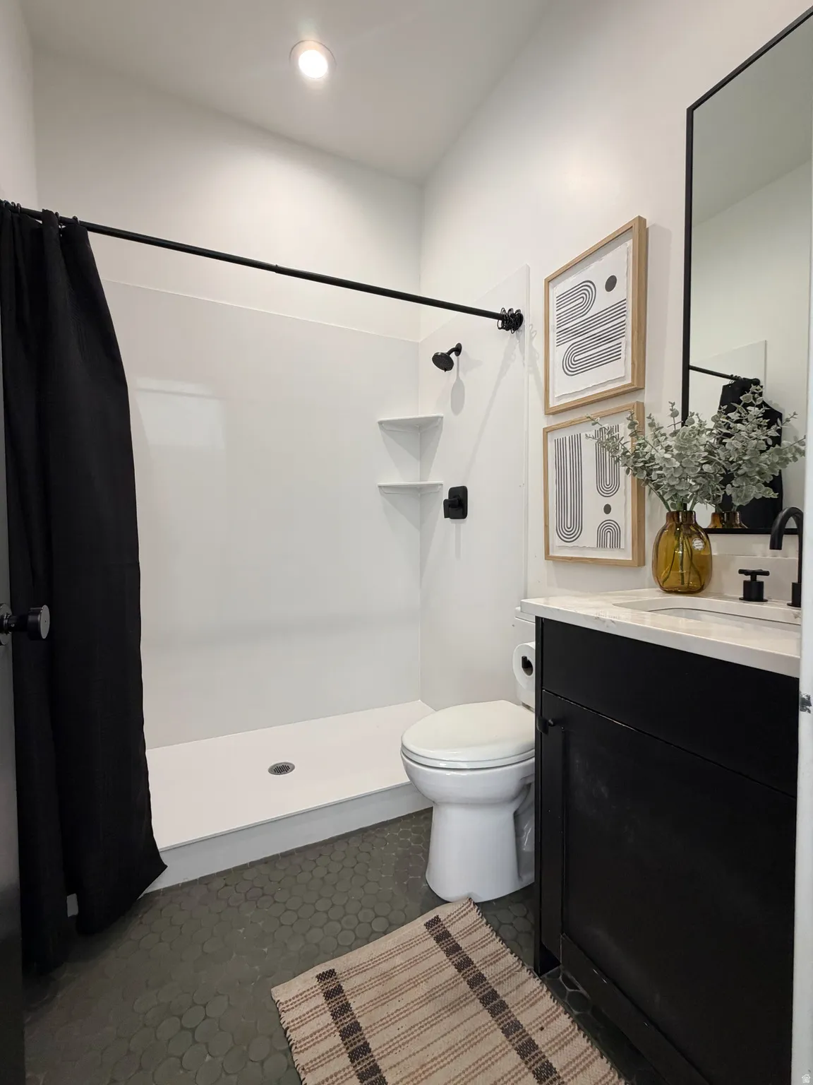 White shower enclosure with integrated shelving, matte black fixtures, dark hexagon tile flooring, and a white countertop vanity with dark cabinetry