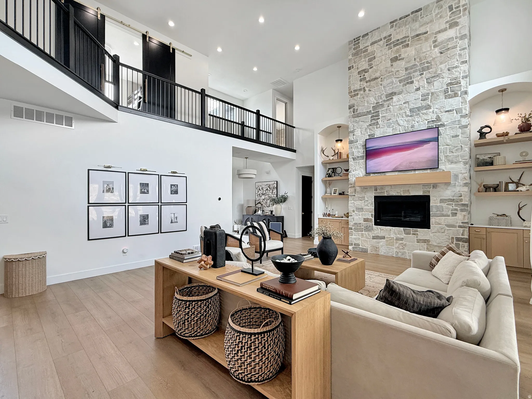 Two-story living area with wood-finish flooring, a stone-clad fireplace, and an upper-level balcony featuring black railings and barn doors