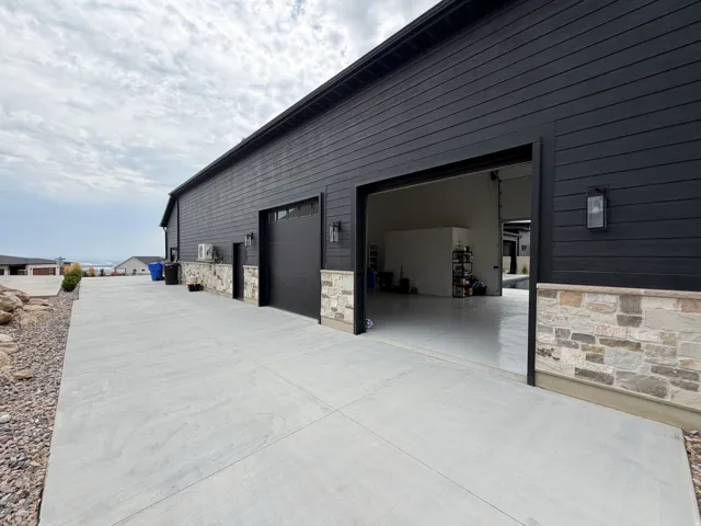 Exterior featuring black horizontal siding, stone veneer accents, two garage doors, and concrete paving