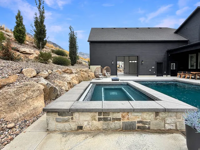 In-ground pool with an integrated spa, stone-clad spa surround, expansive concrete patio, and a dark-toned exterior