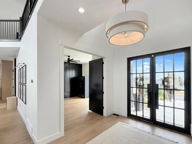 Double entry doors with black frames and glass panels, light wood-finish flooring, a contemporary drum pendant light, white walls, and a black metal staircase railing