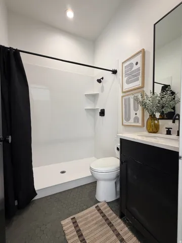 White shower enclosure with integrated shelving, matte black fixtures, dark hexagon tile flooring, and a white countertop vanity with dark cabinetry