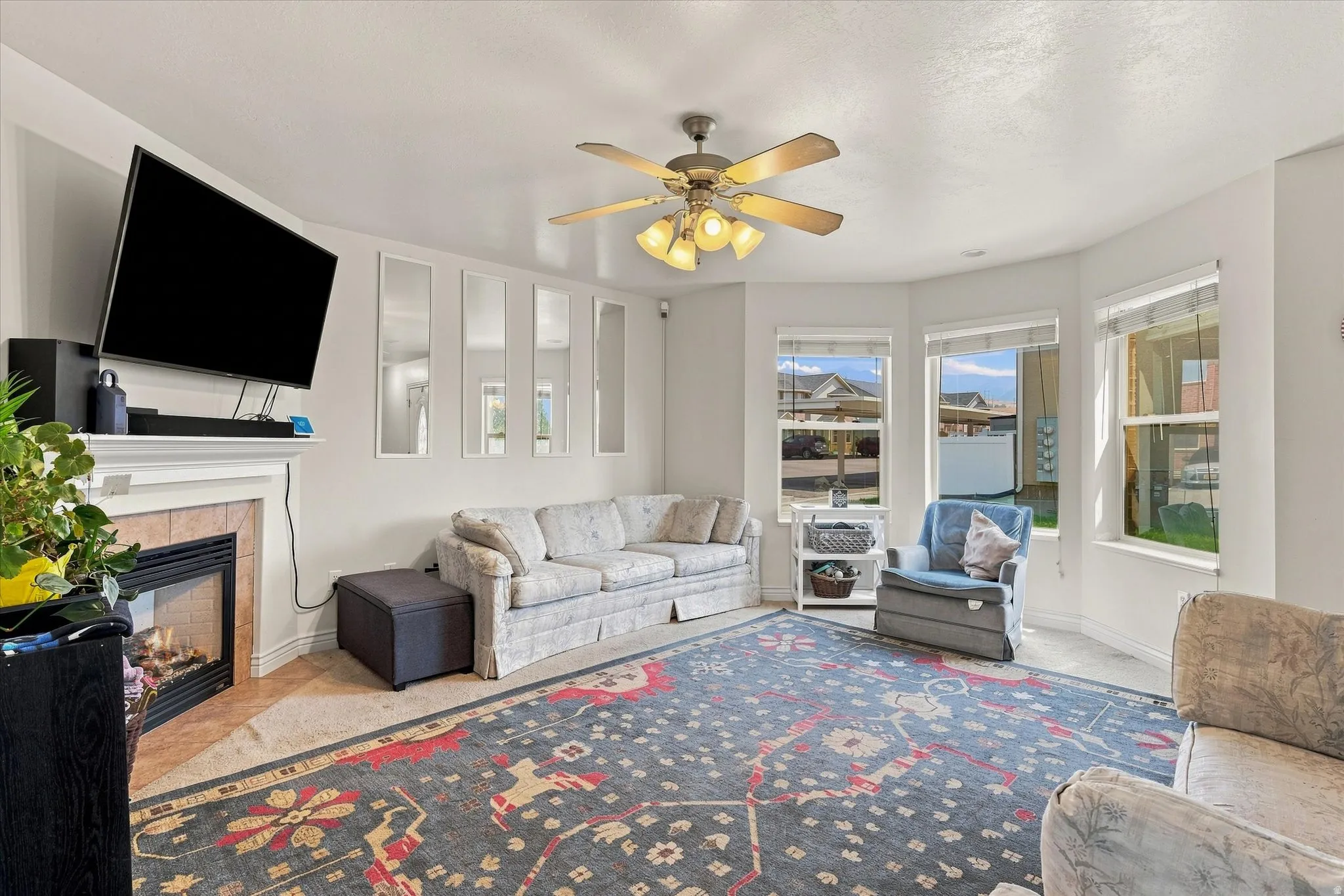 Fireplace with tile surround and mantel, ceiling fan with light fixture, bay windows, and carpet flooring