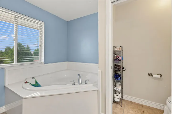 Corner built-in tub with deck-mounted fixtures, large window with horizontal blinds, and tiled flooring