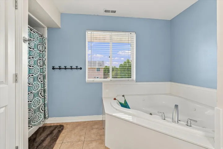 Corner soaking tub with deck-mounted chrome fixtures, adjacent window with blinds, and tile flooring