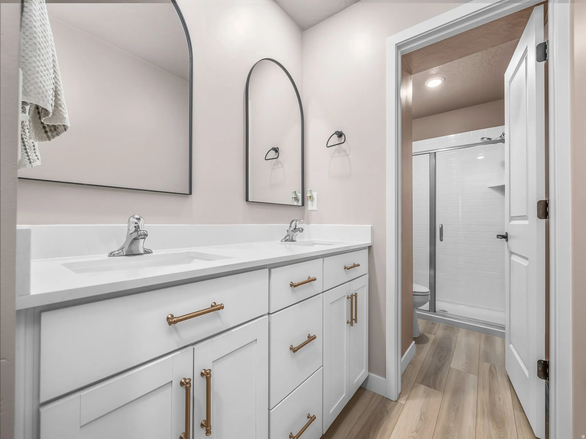 Double vanity with white cabinetry and gold-tone hardware, two integrated sinks with chrome faucets, two arched mirrors, wood-finish flooring, and a shower with a glass enclosure