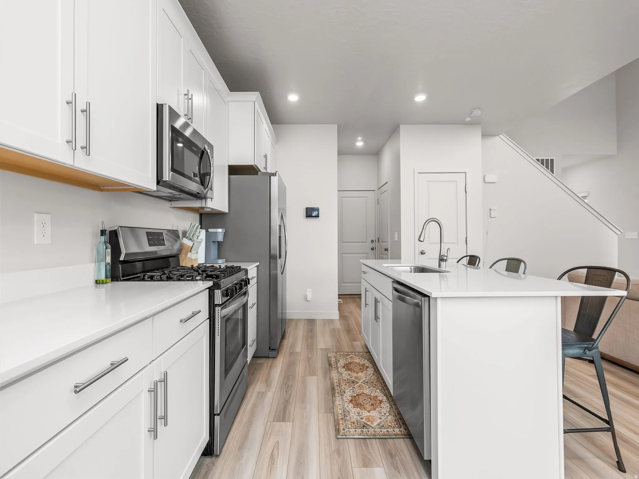 Kitchen featuring white cabinetry, stainless steel appliances, white countertops, a kitchen island with an undermount sink, and wood-finish flooring