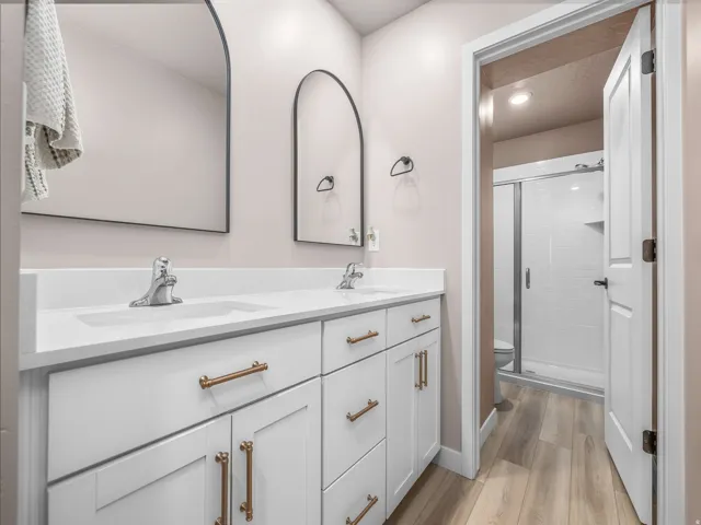 Double vanity with white cabinetry and gold-tone hardware, two integrated sinks with chrome faucets, two arched mirrors, wood-finish flooring, and a shower with a glass enclosure