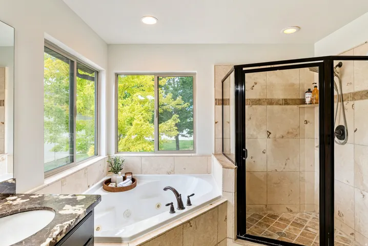 Corner soaking tub, walk-in shower with tile surround, dual windows, recessed lighting, and a vanity with a dark stone countertop
