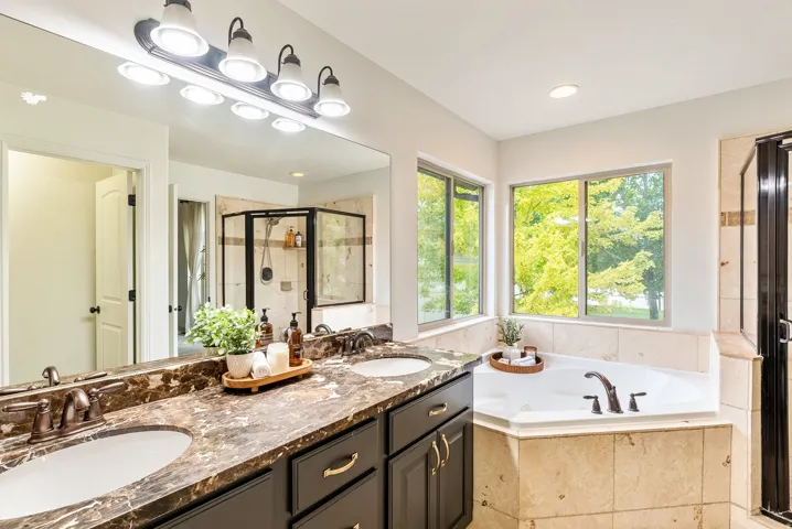 Dual vanity with undermount sinks, large wall-mounted mirror, built-in corner soaking tub, separate shower enclosure, and windows with exterior views