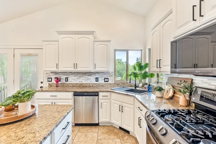 Vaulted ceiling kitchen featuring white cabinetry with black hardware, granite countertops, stainless steel appliances, and a tiled backsplash