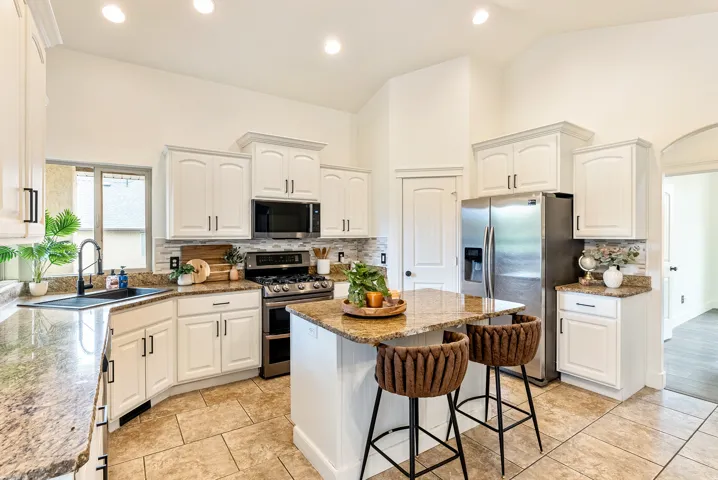 White cabinetry with black hardware, granite countertops, stainless steel appliances, tiled backsplash, and vaulted ceilings