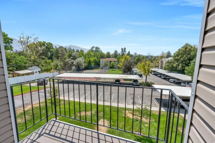 Balcony with black metal railing, overlooking a parking area with covered parking structures, green lawns, and mature trees