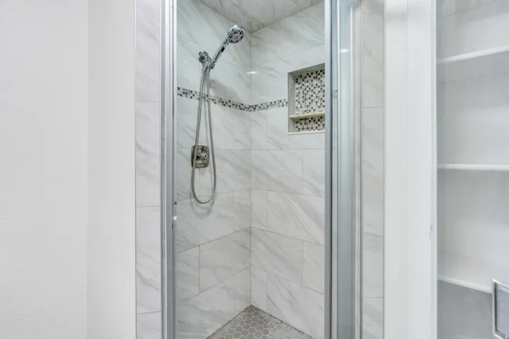 Glass-enclosed shower with white tiled walls, mosaic accent band, built-in tiled niche, and hexagonal floor tiles