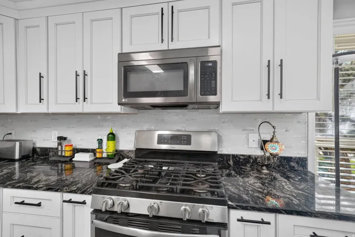 White upper and lower cabinetry with black hardware, stainless steel gas range, coordinating stainless steel microwave, black stone countertops, and white subway tile backsplash