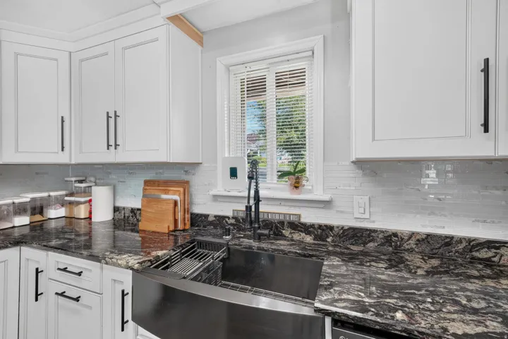 White cabinetry, dark stone countertops, tile backsplash, and a black farmhouse sink