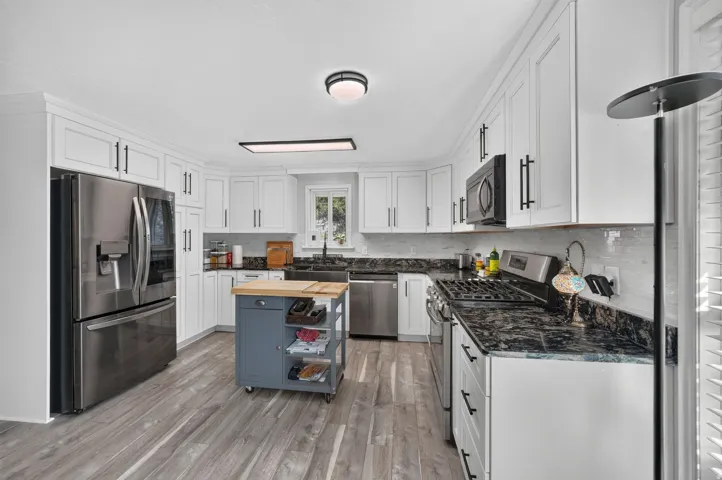 White cabinetry with black hardware, wood-finish flooring, stainless steel appliances, dark countertops, and a tile backsplash
