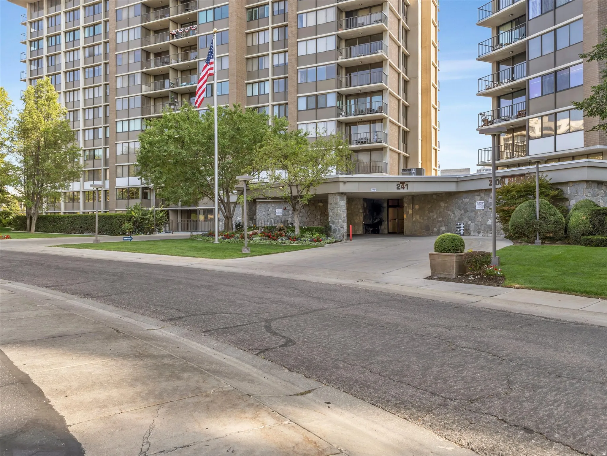 Multi-story building featuring balconies, a covered entry, exterior stone cladding, manicured landscaping, and a paved driveway