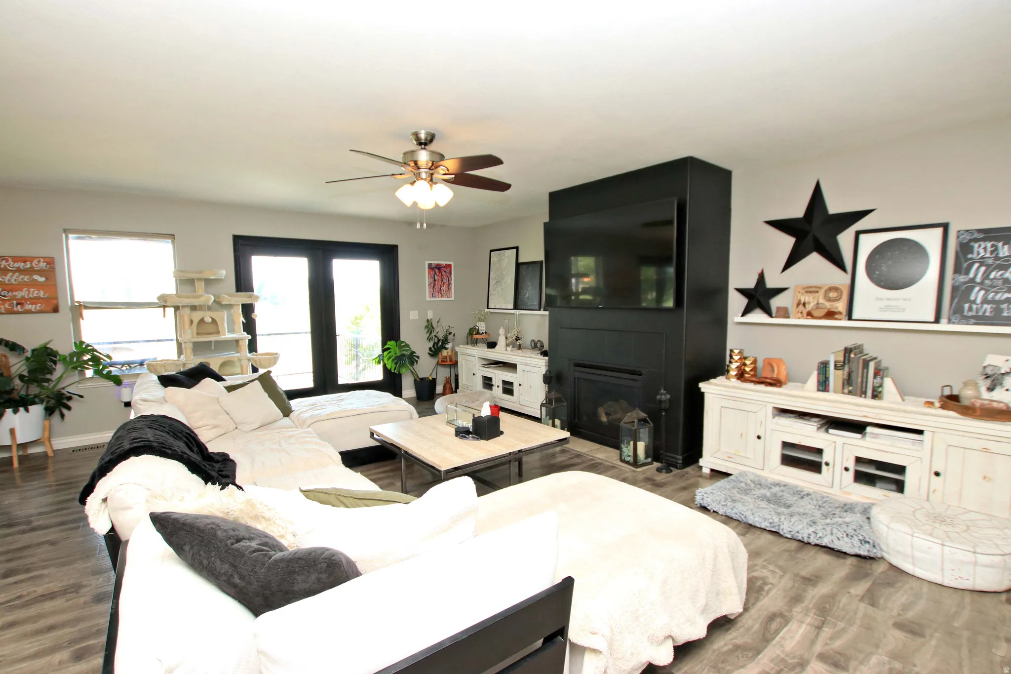 Open-concept living space featuring wood-finish flooring throughout, a ceiling fan with integrated lighting, built-in cabinetry with display shelving, a black accent wall with gas fireplace, and large windows allowing natural light