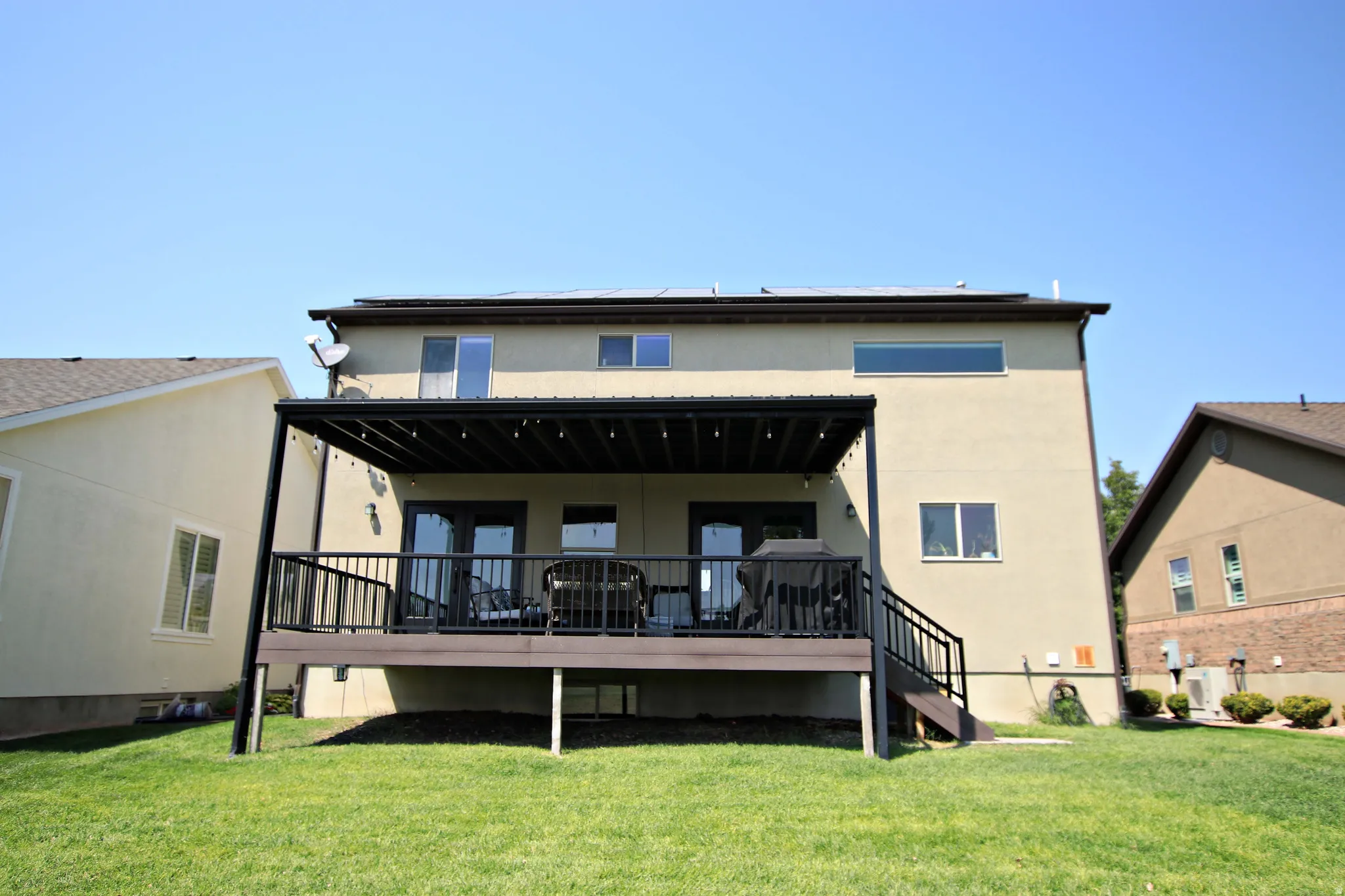 Expansive backyard featuring a manicured lawn, a multi-level deck with black railing, and a covered patio with string lighting
