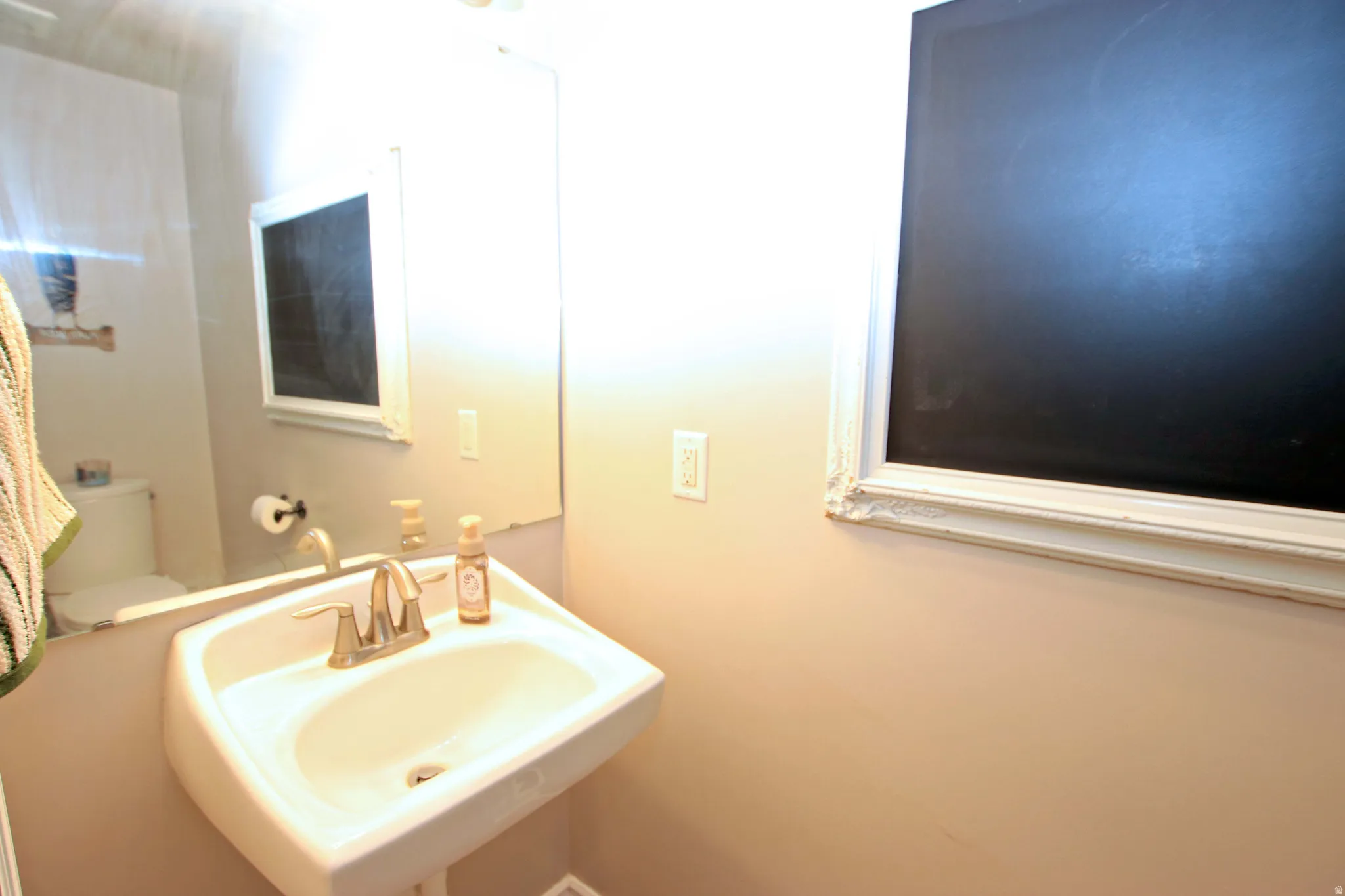 Pedestal sink with a dual-handle faucet, wall-mounted mirror, and a wall-mounted chalkboard with a decorative frame