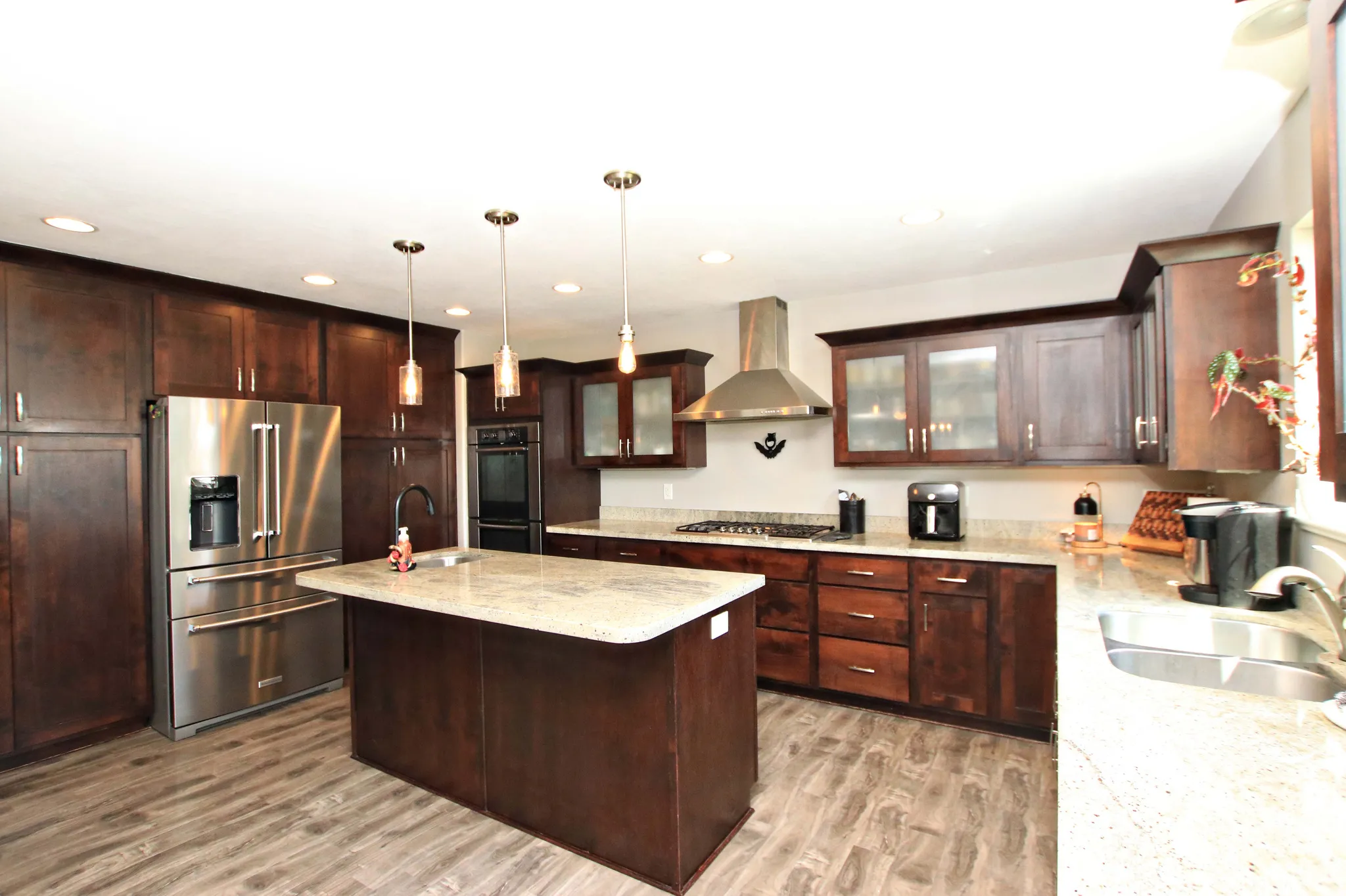Chef's kitchen featuring custom dark wood cabinetry, stainless steel appliances including a multi-door refrigerator and gas cooktop, granite countertops, a large center island with seating, pendant lighting, and wood-finish flooring throughout