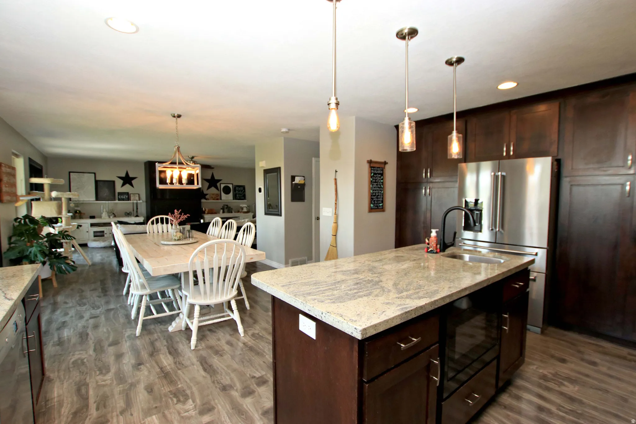 Open-concept kitchen and dining area featuring a large granite topped island with pendant lighting, stainless steel appliances, dark wood cabinetry with brushed nickel hardware, wood-finish flooring, and recessed ceiling lighting throughout the space