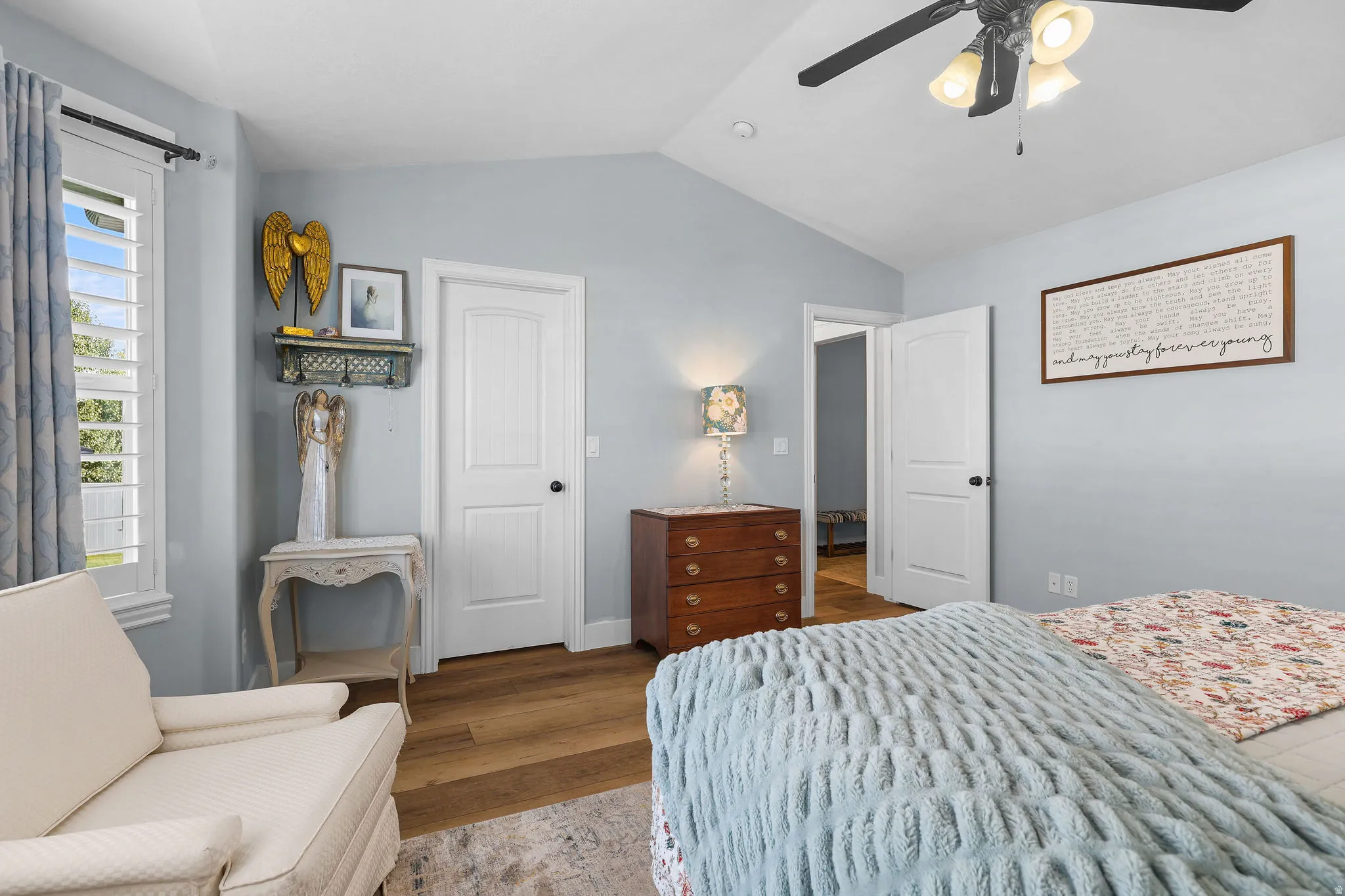Vaulted ceiling, wood-finish flooring, white plantation shutters, ceiling fan with integrated lighting, and a white panel door with black hardware