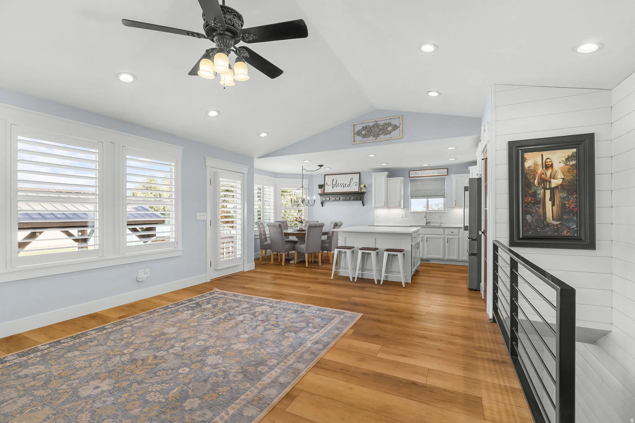 Wood-finish flooring throughout, vaulted ceiling with recessed lighting, white window shutters, kitchen island with seating, and a modern horizontal railing